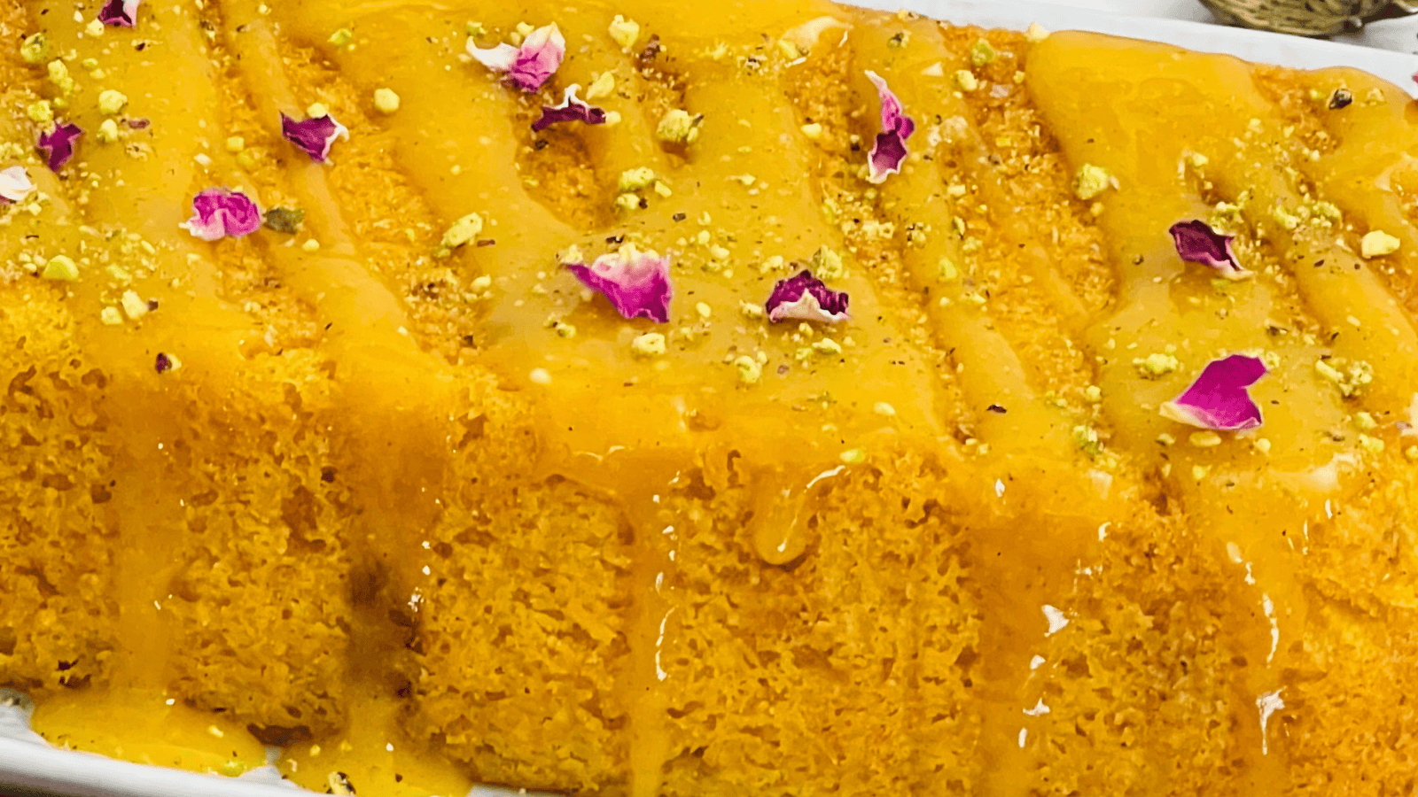 Close-up of a vibrant orange dessert garnished with flower petals and nuts, drizzled with syrup on a white rectangular plate.