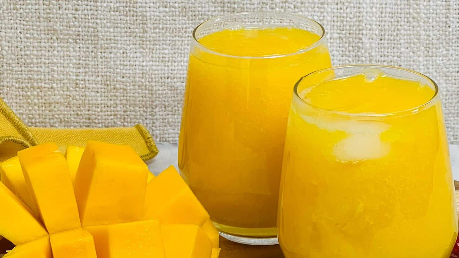 Two clear glasses filled with yellow mango juice sit on a textured surface beside a sliced mango.