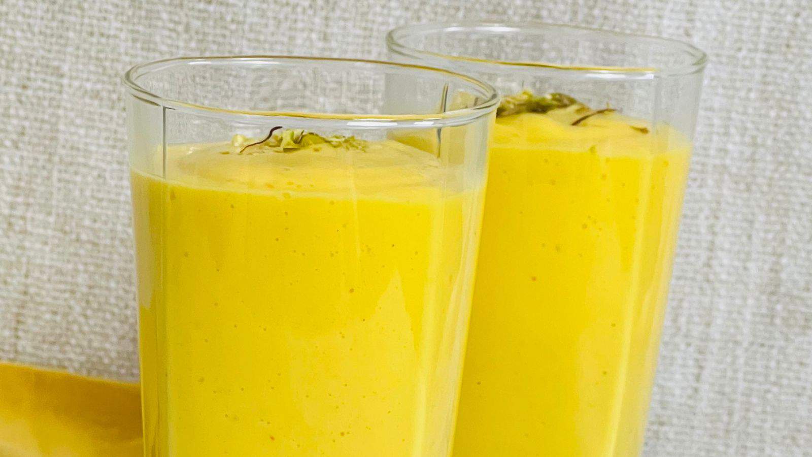 Two tall glasses filled with a thick, yellow mango lassi are placed against a light-colored textured background.