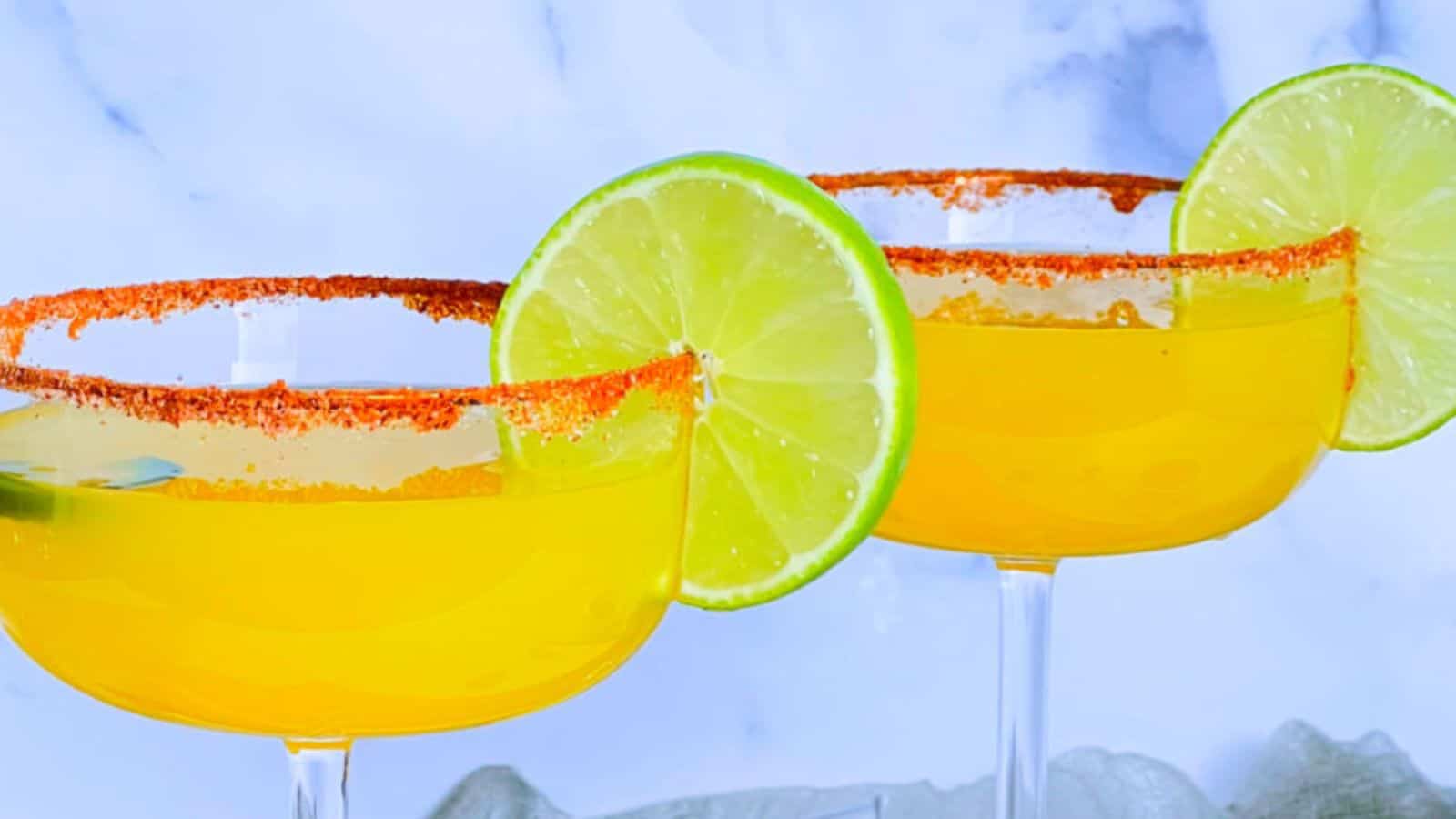 Two yellow cocktails served in wide-rimmed glasses, each garnished with a slice of lime and a salted, spicy rim, are set against a light, marble-like background.