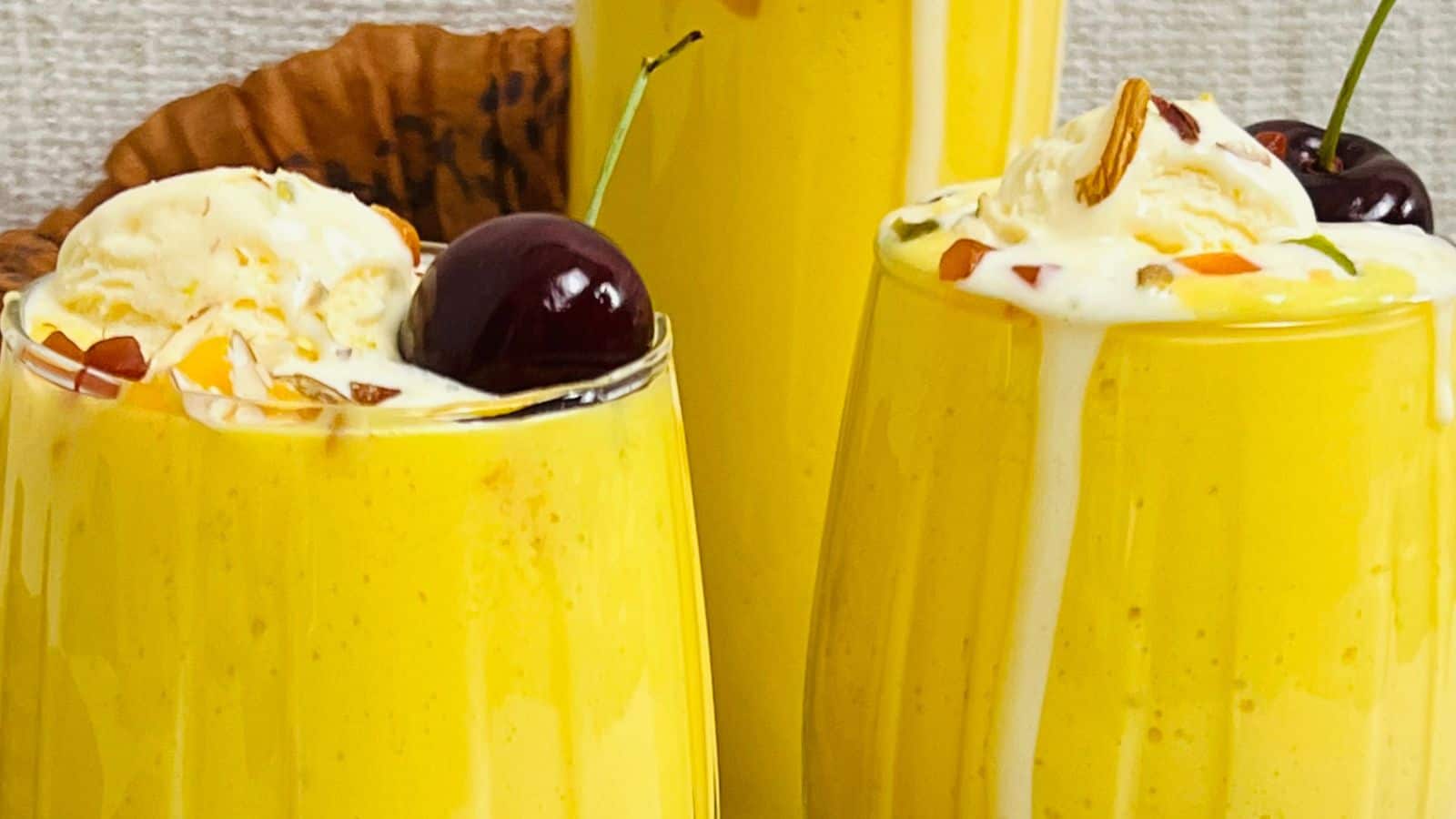 Two glasses of mango lassi topped with vanilla ice cream, cherries, and nuts. A large pitcher of mango lassi is visible in the background.