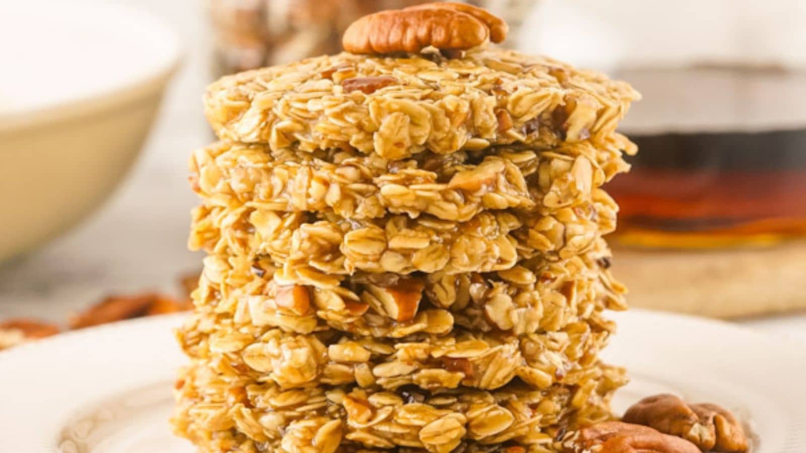 A stack of no-bake maple pecan cookies on a white plate, with syrup and a bowl in the background.