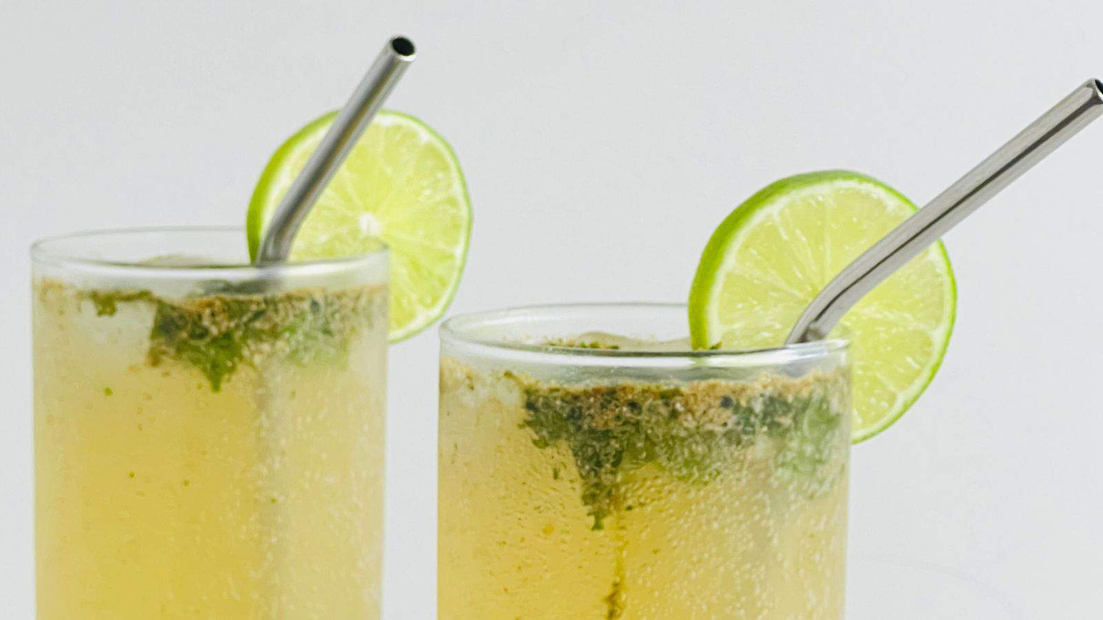 Two glasses of a yellow drink garnished with lime slices and metal straws. The drink appears to have herbs or spices sprinkled on top.