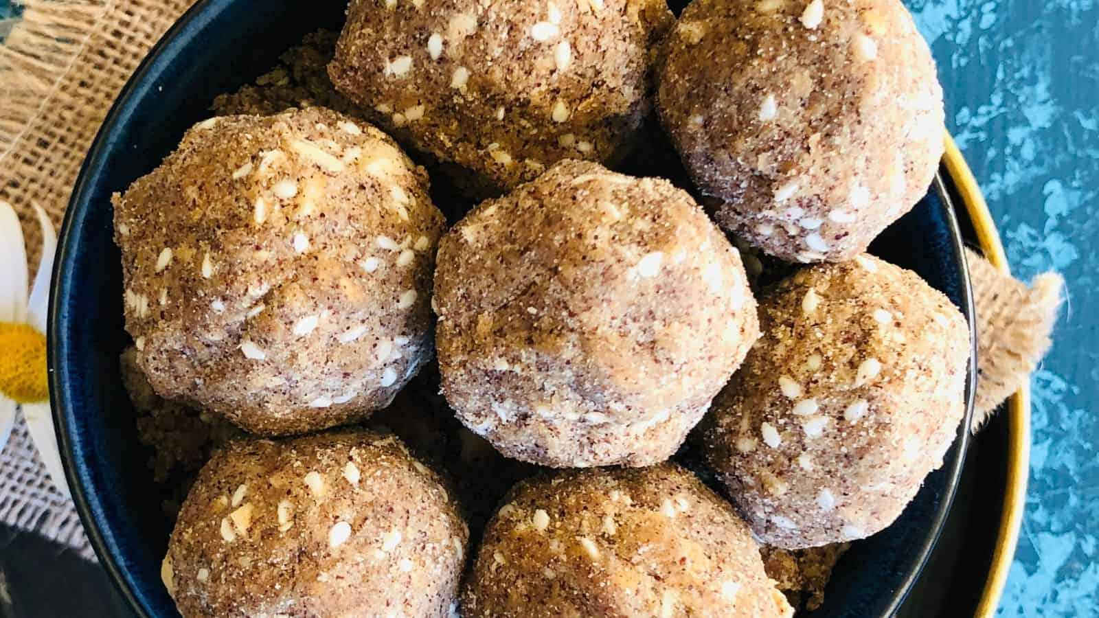 A black bowl filled with round, brown-colored dessert balls featuring small white seeds sits on a rustic textured cloth.