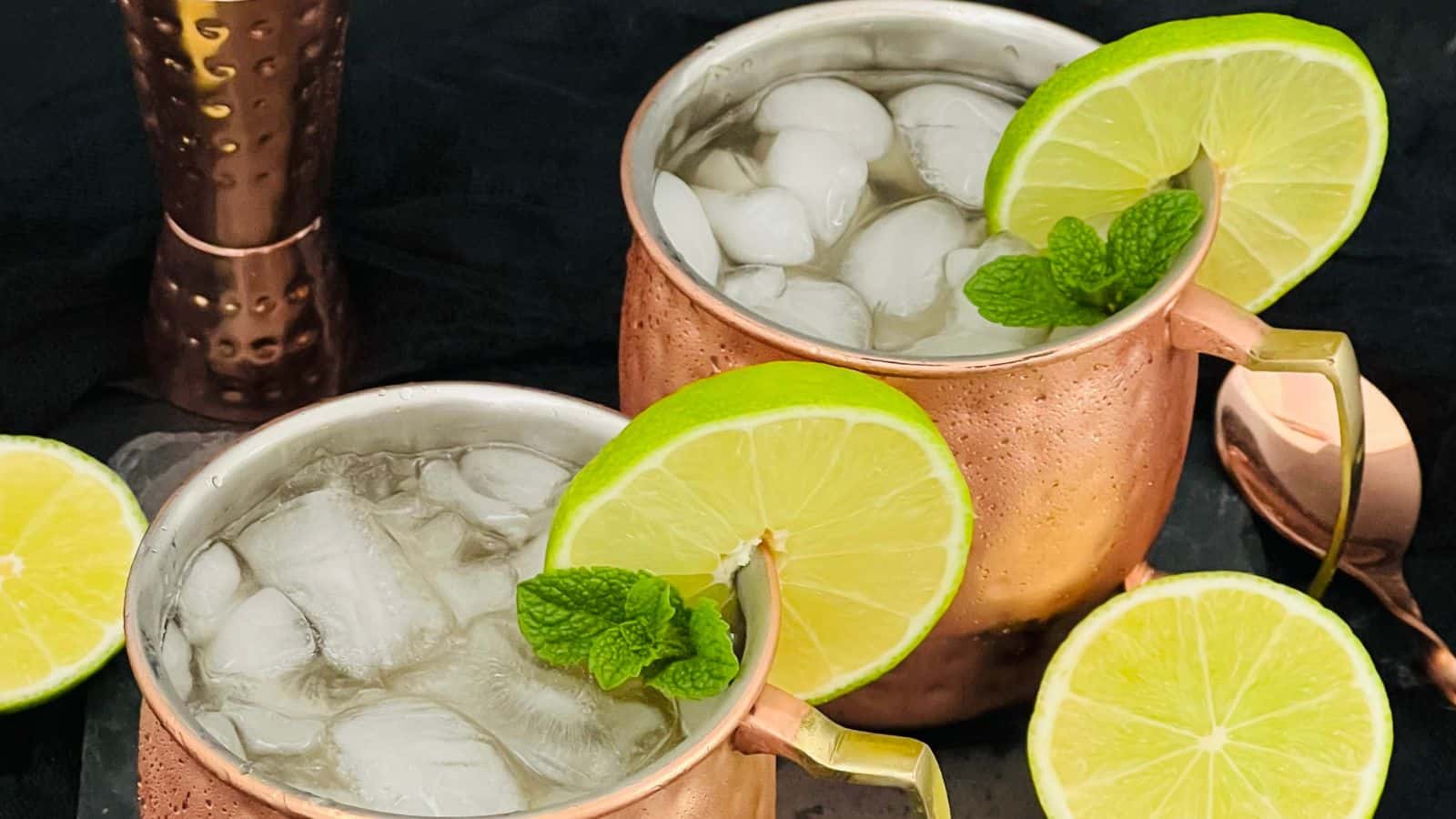 Two copper mugs filled with ice and a clear beverage, each garnished with a slice of lime and a sprig of mint. Slices of lime are placed beside the mugs. A copper item is in the background.
