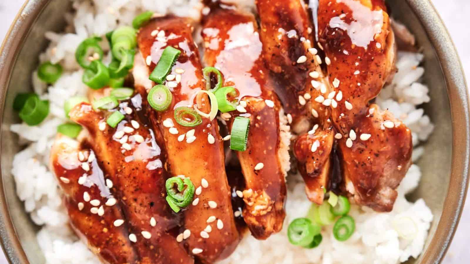 A bowl of white rice topped with glazed chicken and garnished with green onions and sesame seeds.