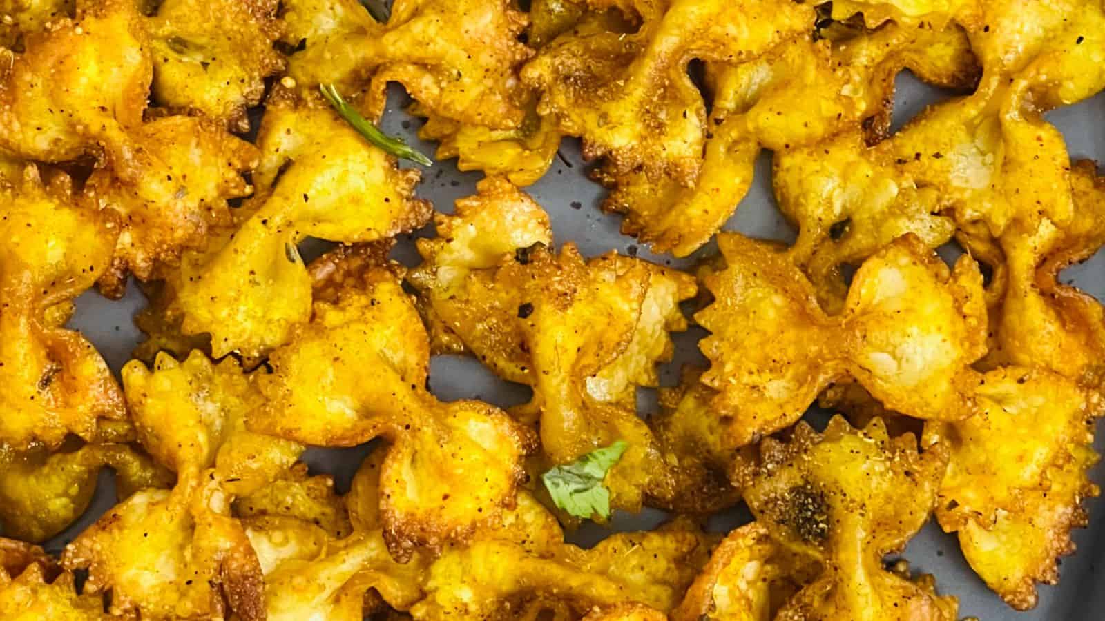 A close-up view of crispy, seasoned bow-tie pasta on a pan, golden-brown and garnished with small green herb pieces.