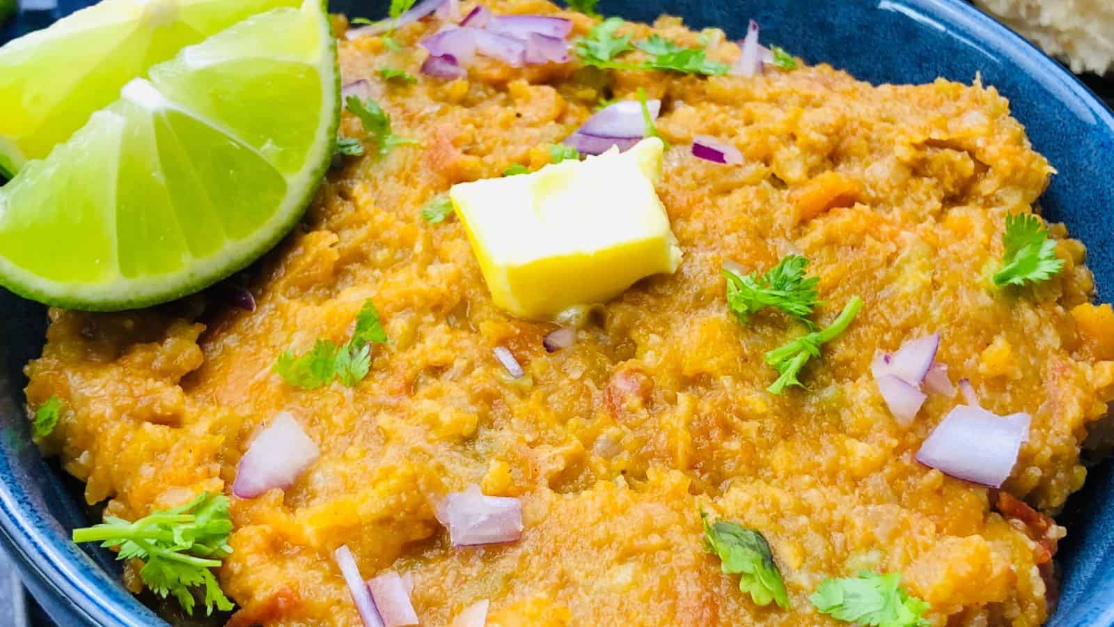 A close-up of a bowl of mashed vegetables garnished with red onions, cilantro, a pat of butter, and two lime wedges.