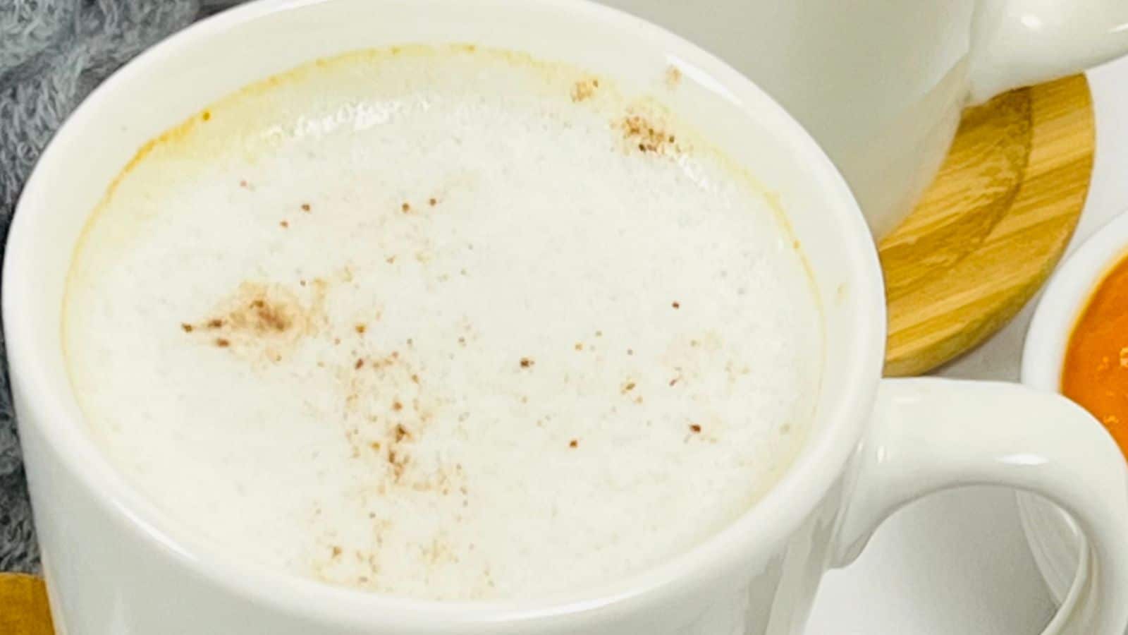 A close-up image of a white ceramic mug filled with a frothy beverage, sprinkled with cocoa powder on top. The mug is placed on a wooden coaster next to an orange sauce in a small container.