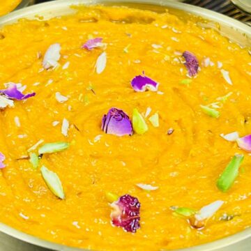 A bowl of orange-colored dessert garnished with flower petals and nuts, placed on a metallic surface.