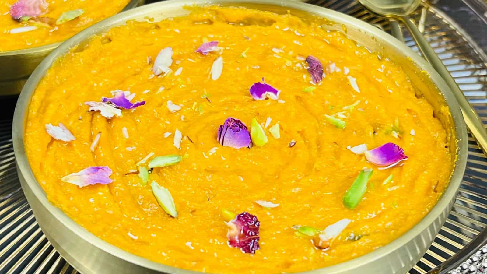 A bowl of orange-colored dessert garnished with flower petals and nuts, placed on a metallic surface.