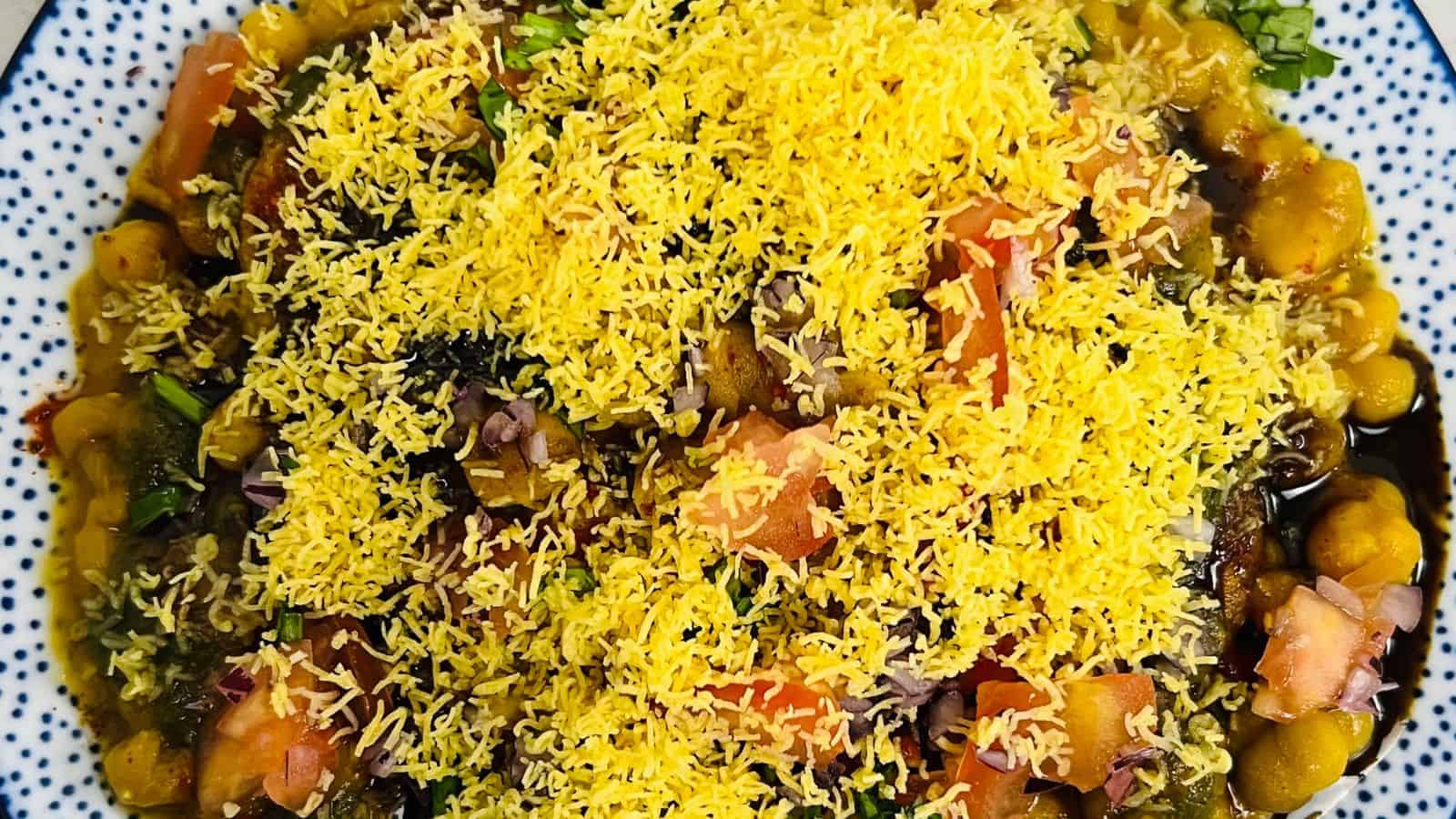 A close-up of a dish consisting of assorted vegetables and pulses, topped with a generous layer of yellow sev (crispy noodles).