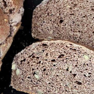 Sliced loaf of whole grain bread with two separate slices placed next to it on a dark background. The bread has a textured, crusty exterior and contains visible seeds.