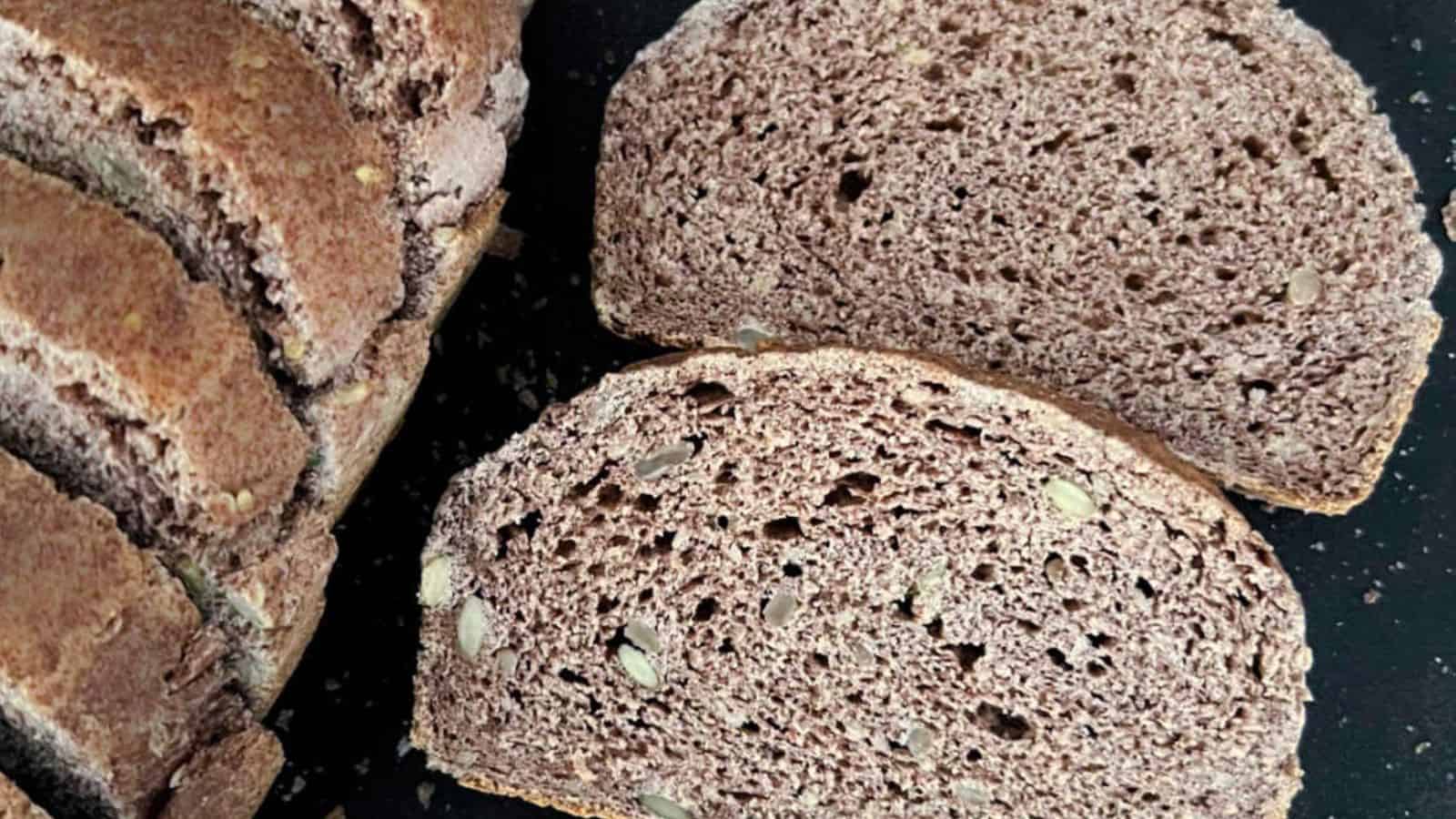 Sliced loaf of whole grain bread with two separate slices placed next to it on a dark background. The bread has a textured, crusty exterior and contains visible seeds.