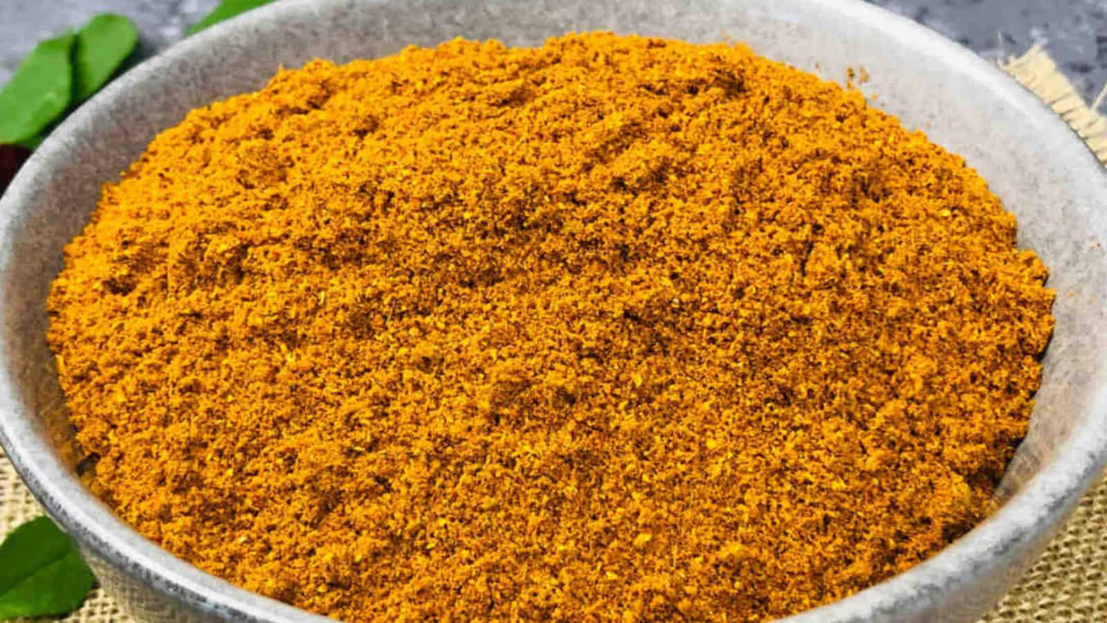 A bowl filled with a large amount of ground yellowish-brown spice, likely a type of curry powder, placed on a light-colored surface.