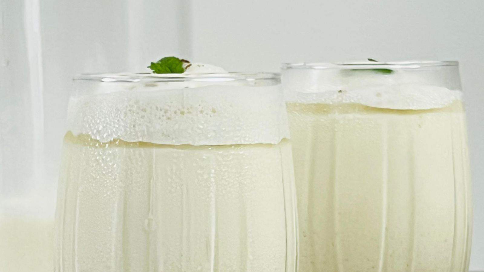 Two frothy glasses of a creamy white beverage, possibly a milkshake or smoothie, garnished with a mint leaf on top.