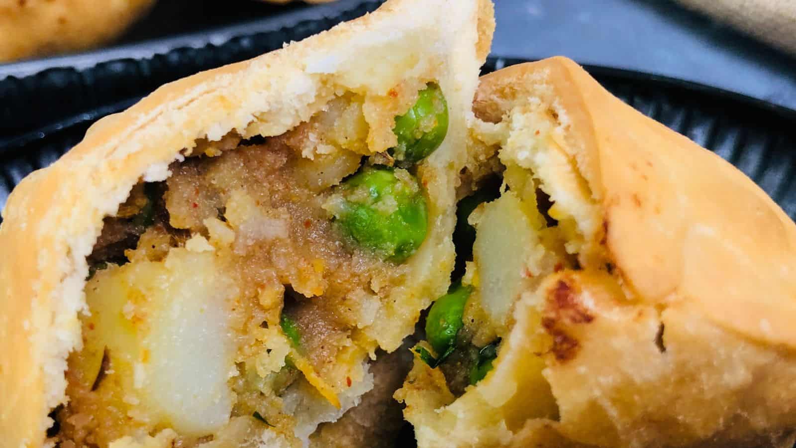 Close-up of a cut samosa revealing its filling of potatoes, peas, and spices. The golden-brown crust appears crispy, and the stuffing is well-cooked. The background includes more samosas and a black plate.