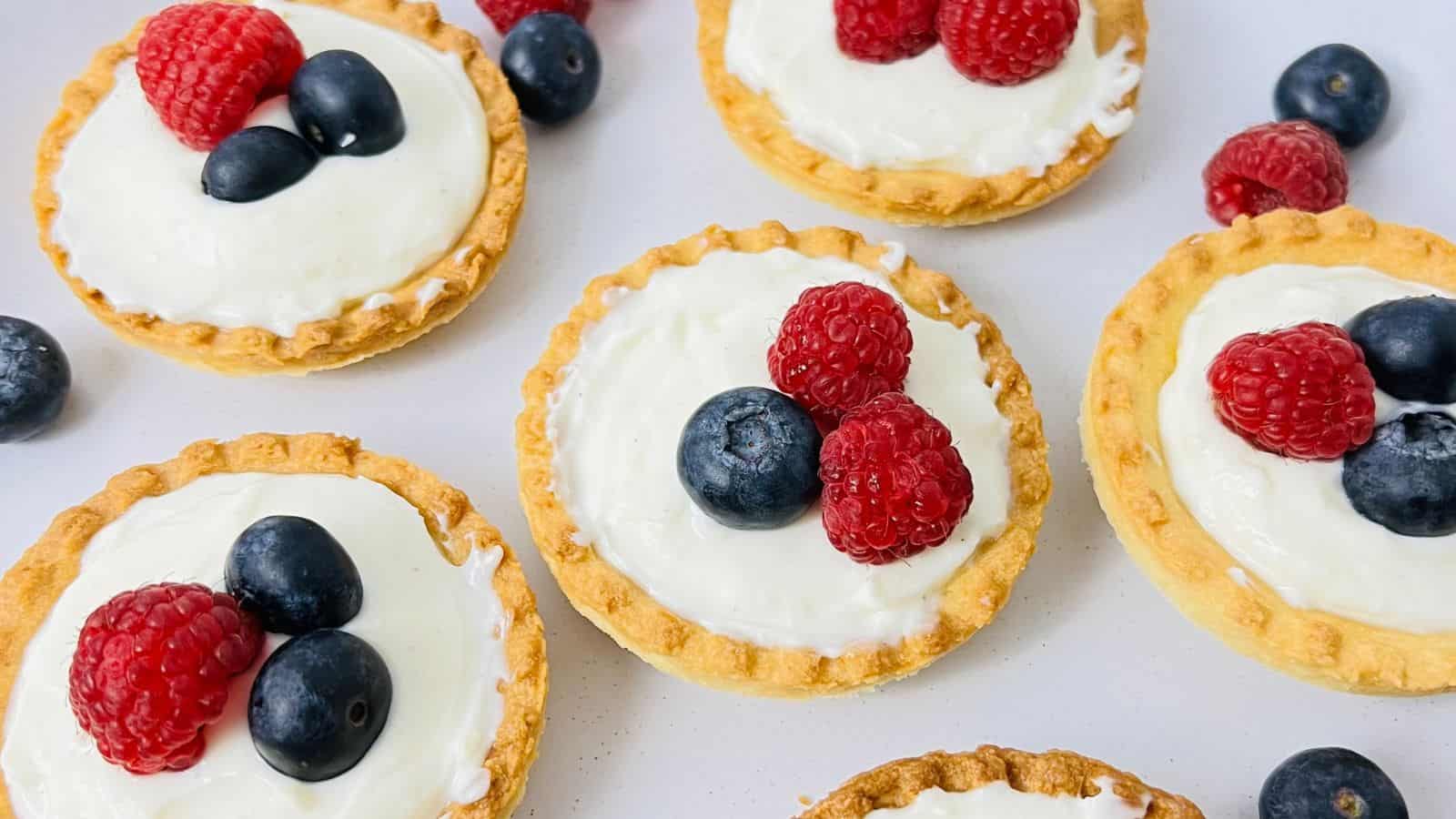 Mini tartlets with creamy filling, topped with fresh raspberries and blueberries, arranged on a white surface.