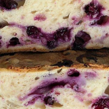 Close-up of two slices of freshly baked bread with visible blueberries and swirls of blueberry juice within the soft crumb. The crust appears lightly browned.