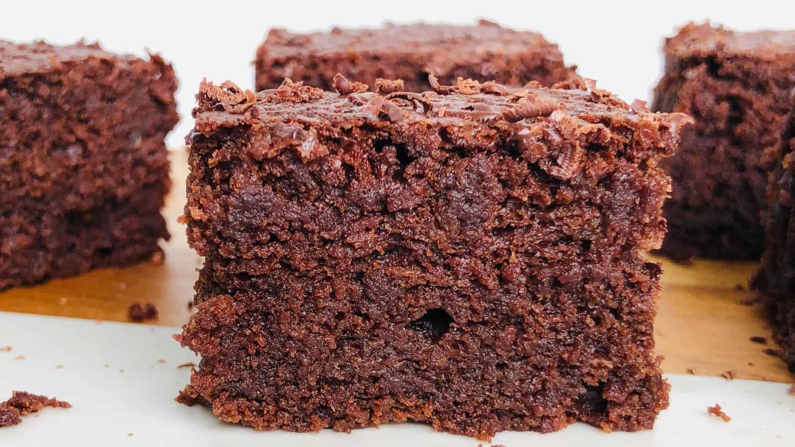Close-up of a square cut chocolate brownie with a rich, dense texture, resting on a light-colored surface. More brownies are visible in the background.