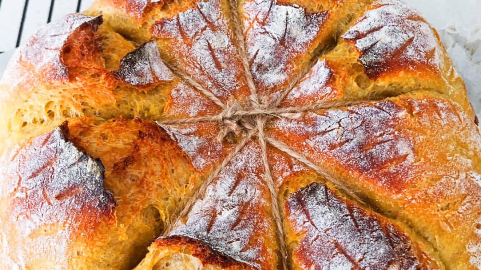 Close-up of a round, crusty loaf of bread with a decorative top and dusting of flour. The bread has deep, golden-brown ridges and a visible twine around its center.