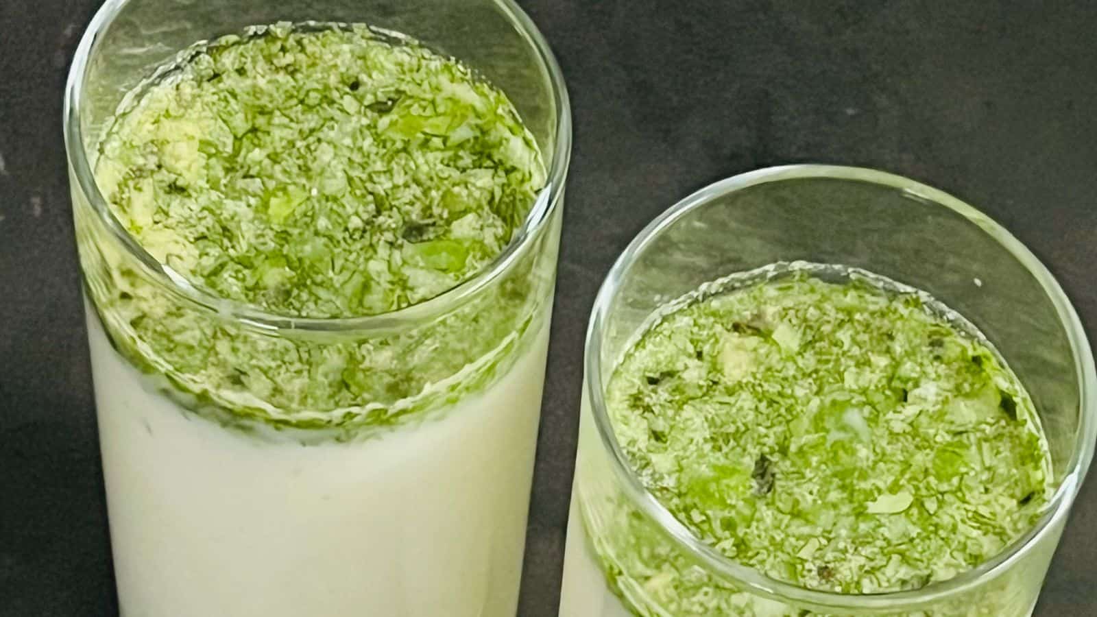 Two glasses filled with a white liquid topped with green herbs on a dark surface.