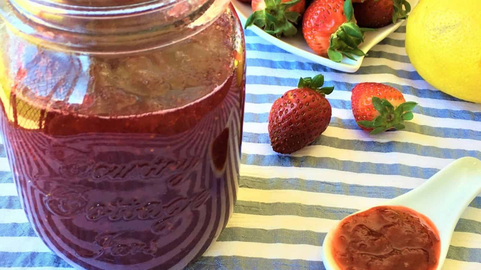 A jar of strawberry jam next to whole strawberries, a white dish with a spoonful of jam, and a lemon, all placed on a blue and white striped cloth.