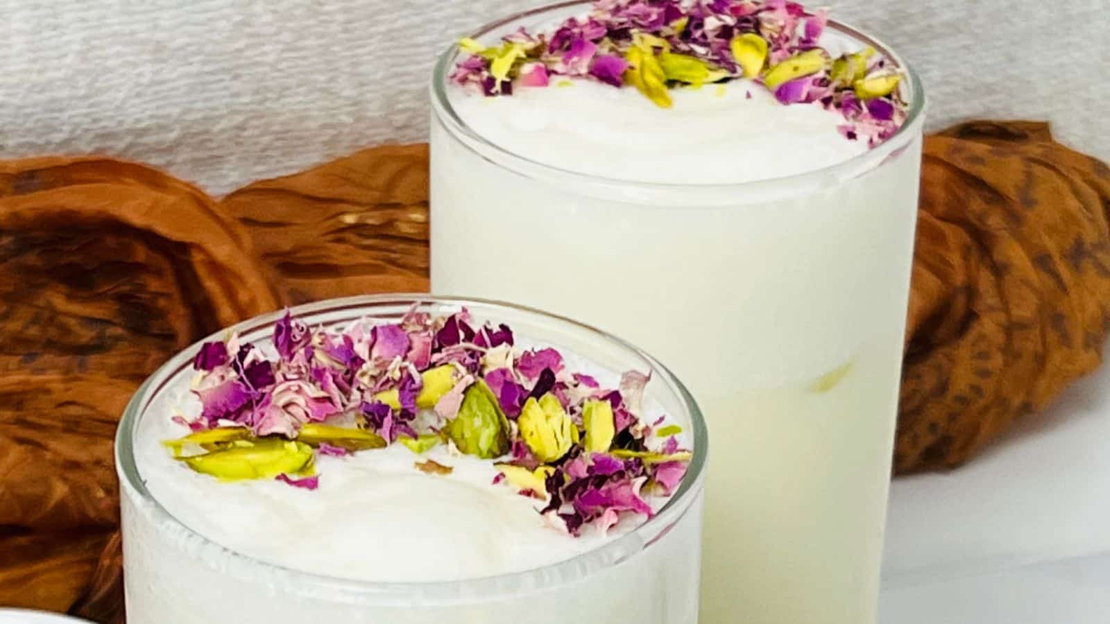 Two glasses of creamy white beverage garnished with chopped pistachios and purple flower petals, placed on a white surface with a textured brown element in the background.