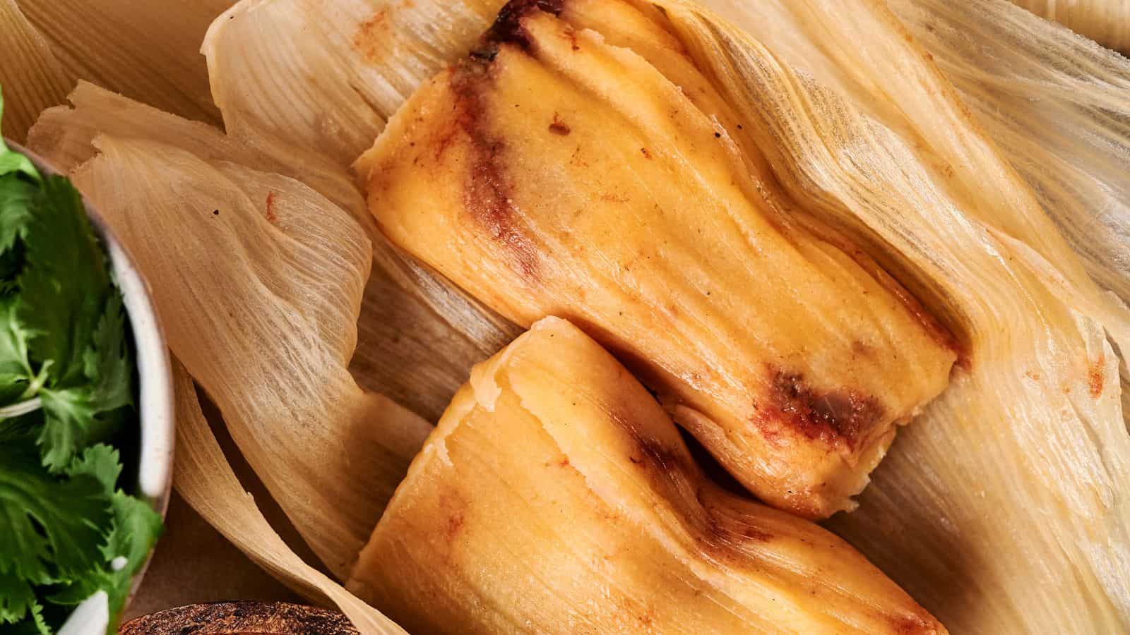 Two cooked tamales nestled in corn husks, with a bowl of fresh cilantro partially visible on the left side.