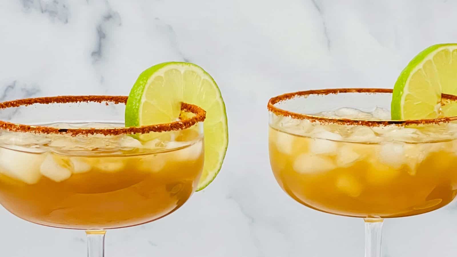 Two cold cocktails in stemmed glasses are garnished with lime slices and rimmed with chili powder. The background is a white marble surface.