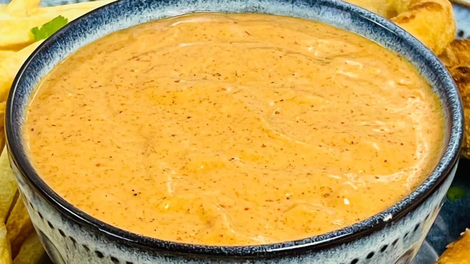 A close-up of a bowl filled with a creamy, orange-colored dipping sauce. The bowl has a blue rim, and the sauce has visible specks of spices.