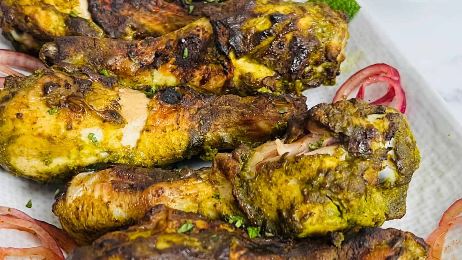 Close-up of several grilled chicken drumsticks coated with a yellowish-green seasoning, garnished with herbs and served on a white plate. Sliced red onions are placed alongside the chicken.