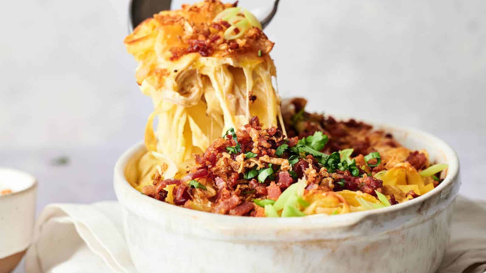 Close-up of a casserole dish with a cheesy pasta bake, topped with crispy bacon bits and green onions. A serving spoon is lifting a portion, showing melted cheese strands.