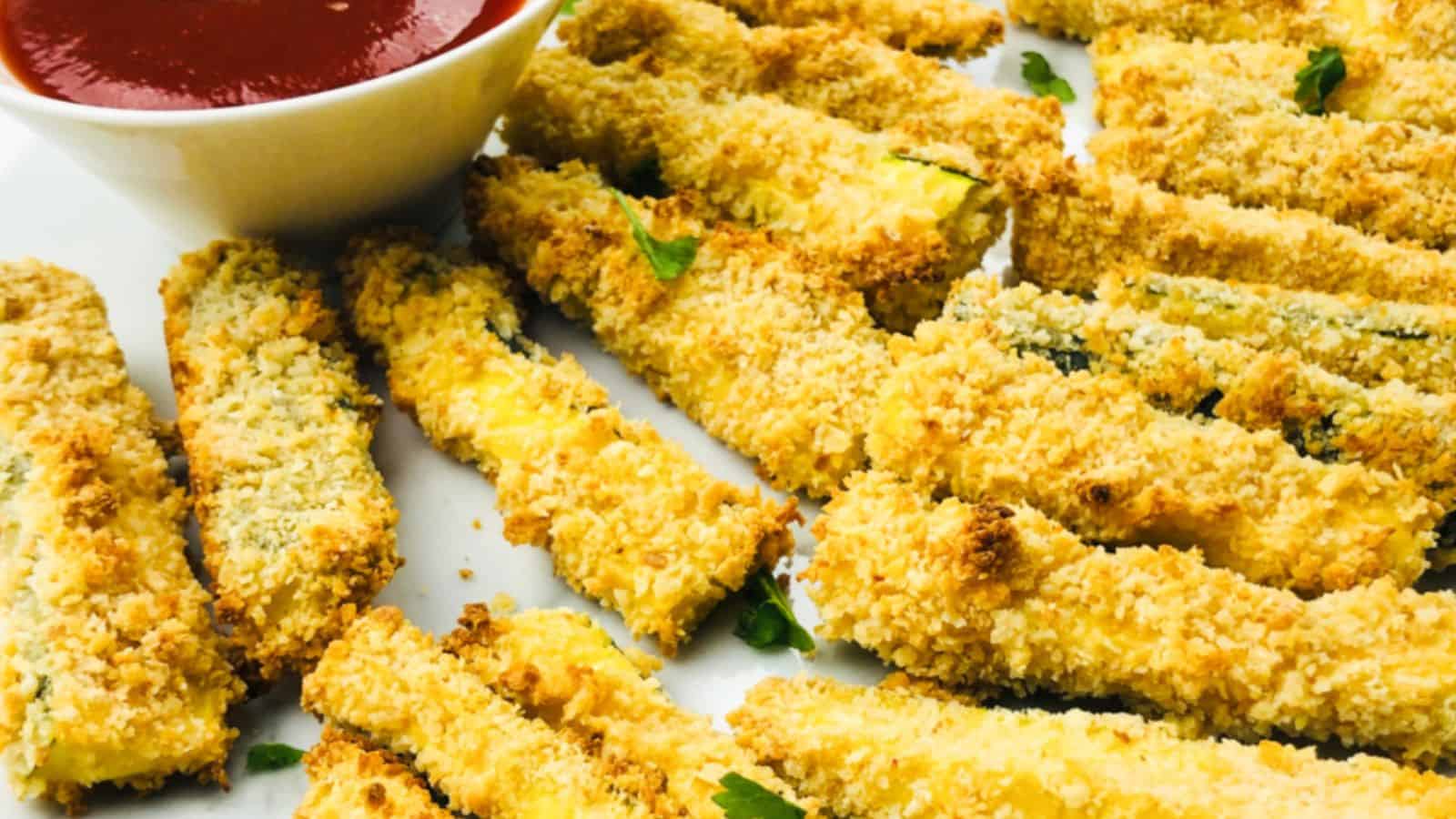 A plate of breaded and baked zucchini sticks is served with a small bowl of ketchup.