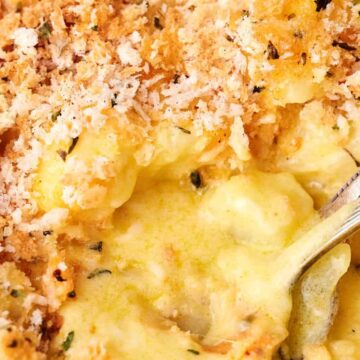 Close-up of a creamy, golden-brown macaroni and cheese dish with a crunchy breadcrumb topping reminiscent of comforting funeral potatoes. A spoon is partially submerged in the cheese sauce.