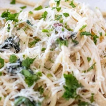 A close-up of a plate of Creamy Tuscan Pasta topped with a rich white sauce, grated cheese, and garnished with chopped parsley.