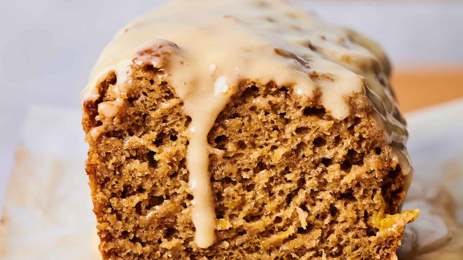 A close-up of a sliced loaf of pumpkin bread with a light tan glaze dripped over the top. The bread has a moist and textured look, with visible air bubbles throughout.